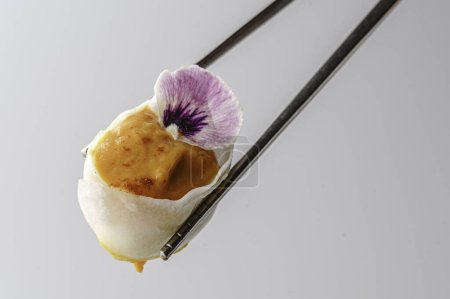 Téléchargez les photos : Une délicate boulette de dim sum remplie de contenu salé, délicatement tenue par une paire de baguettes, est ornée d'une fleur pourpre comestible, créant une élégante fusion de cuisine et d'art. - en image libre de droit