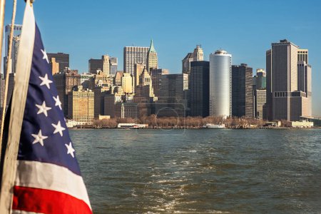 Téléchargez les photos : Un drapeau américain flotte au premier plan avec l'horizon du Lower Manhattan comme toile de fond, vu de l'eau. - en image libre de droit