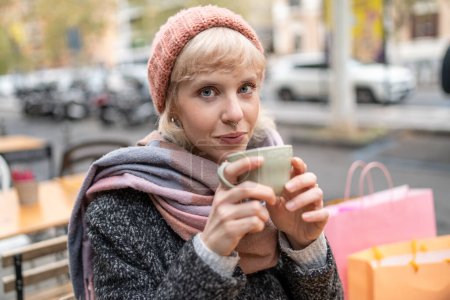Téléchargez les photos : Une jeune femme blonde profite d'une tasse de café chaude dans une rue de la ville, incarnant une journée d'hiver détendue. - en image libre de droit