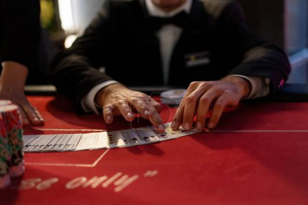 Téléchargez les photos : Gros plan des mains d'un marchand de cartes distribuant habilement des cartes sur une table de casino rouge vibrante. - en image libre de droit
