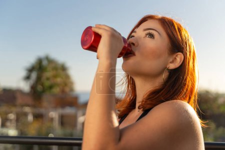 Téléchargez les photos : Une rousse en forme boit dans une bouteille rouge, restant hydratée pendant une séance d'entraînement en plein air. - en image libre de droit
