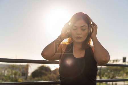 A contemplative young woman enjoys a peaceful moment, listening through her headphones as the sun sets, enveloping her in a warm glow.