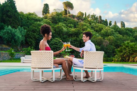 Couple enjoying a sunny day with a toast by a luxurious pool, surrounded by lush greenery.