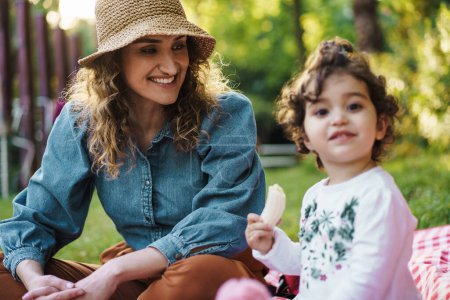 Photo for A smiling mother in a hat shares a joyful moment with her curly-haired daughter in a park. - Royalty Free Image