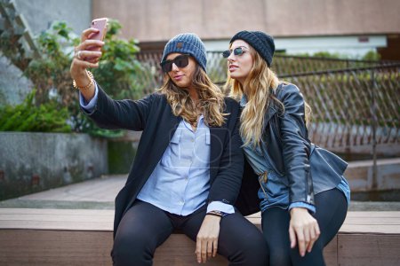 Photo for Two fashionable young women in casual wear and beanies taking a selfie with a smartphone while sitting outside, showcasing friendship and modern lifestyle. - Royalty Free Image