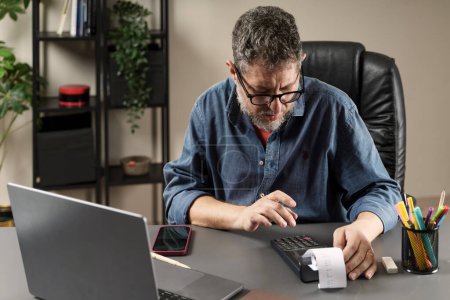 Photo for Concentrated man using a calculator to manage finances beside a laptop, exemplifying meticulous financial planning. - Royalty Free Image