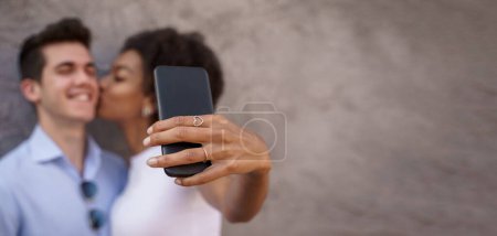 Foto de Pareja multicultural compartiendo un momento romántico mientras se toma una selfie con un teléfono inteligente sobre un fondo texturizado. Concéntrate en el teléfono, caras borrosas. - Imagen libre de derechos