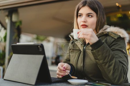 Photo for A focused young curvy woman in a green jacket enjoys a warm cup of coffee as she works on her tablet, embodying the flexibility of remote work. - Royalty Free Image
