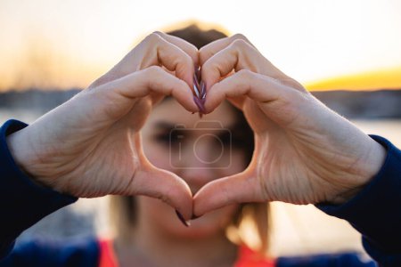 Photo for A young woman creates a heart shape with her hands, capturing a moment of affection against the warm glow of the setting sun. - Royalty Free Image