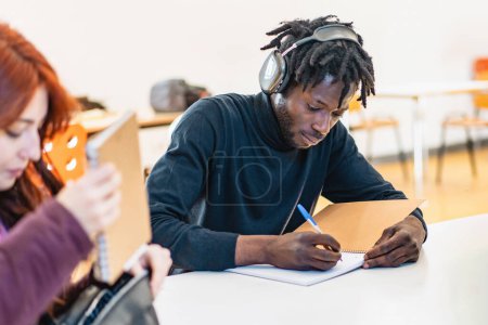 Foto de Joven estudiante africano con auriculares escribiendo intensamente notas en un escritorio de clase, absorbido en el estudio. - Imagen libre de derechos