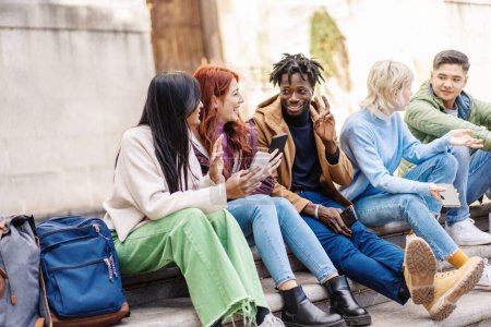 Téléchargez les photos : Divers amis de la génération Z partagent des rires et du contenu sur les téléphones assis ensemble à l'extérieur. - en image libre de droit