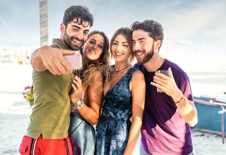 Téléchargez les photos : Groupe d'amis joyeux prenant un selfie avec des boissons sur une plage ensoleillée. - en image libre de droit