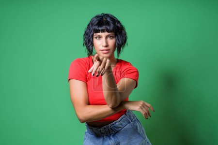 Téléchargez les photos : Femme brune confiante dans une chemise rouge avec une coiffure à la mode posant pointant sur vous sur un fond d'écran vert - Style urbain et concept de mode - en image libre de droit