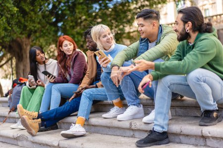 Diverse friends share and laugh over content on smartphones, sitting on steps in an urban setting.