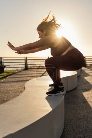 Téléchargez les photos : Femme active dans un squat profond contre le lever du soleil - Adopter un mode de vie sain avec des séances d'entraînement le matin en ville. - en image libre de droit