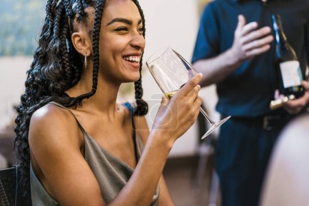 Téléchargez les photos : Jeune femme aux cheveux tressés souriant tout en tenant un verre de vin blanc - concept de plaisir et de style de vie lors d'une dégustation de vin. - en image libre de droit