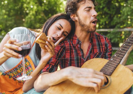 Téléchargez les photos : Femme dégustant un sandwich et du vin serenade par un homme jouant de la guitare - Pique-nique en plein air décontracté avec musique live - en image libre de droit