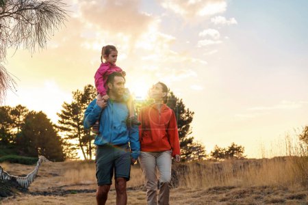 Téléchargez les photos : Jeune famille en randonnée décontractée, avec enfant sur les épaules du père, profitant du magnifique coucher de soleil dans la nature. - en image libre de droit