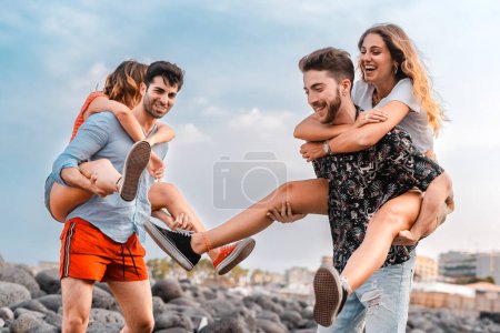 Photo for Playful couple on vacation - A vibrant scene of two young couples engaging in playful piggyback rides along the rocky beach, with laughter filling the air and a sense of carefree youthfulness. - Royalty Free Image