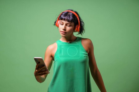 Téléchargez les photos : Femme en robe verte écoute de la musique avec des écouteurs orange, tenant le téléphone style décontracté répond à la commodité technique. - en image libre de droit