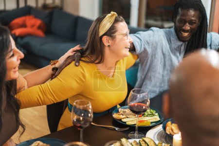 Téléchargez les photos : Un groupe diversifié d'amis partagent un repas joyeux lors d'un dîner confortable à la maison, entouré de rire et de bonne nourriture - dîner maison décontractée multiethnique avec du vin. - en image libre de droit