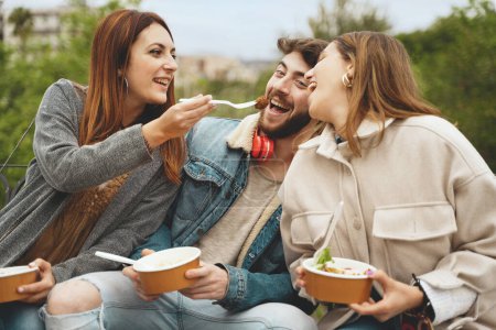 Téléchargez les photos : Trois amis partageant un repas sain ensemble à l'extérieur, souriant et appréciant la compagnie de l'autre - concept de salle à manger extérieure décontractée - en image libre de droit