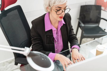 Elegante mujer mayor que trabaja en la computadora de escritorio - ambiente de oficina, profesionalismo - Experiencia, lugar de trabajo moderno.