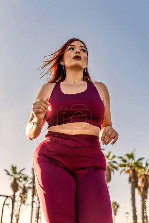 Plus-Size-Frau läuft im Freien in kastanienbrauner Sportbekleidung und zeigt Entschlossenheit und Fitness-Motivation, gesunden Lebensstil, hellen und sonnigen Tag, aktive und dynamische Bewegung