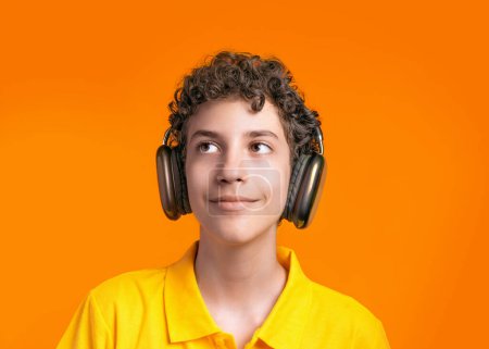 Nachdenklicher Junge mit lockigem Haar, Kopfhörer und gelbem Hemd, leuchtend orangefarbenem Hintergrund, lächelnd aufblickend, ausdrucksstarkes und kreatives Porträt, jugendliche Energie und Phantasie