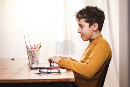 Young boy sits at a desk, using a laptop for online learning. School supplies and natural sunlight - focused and educational atmosphere at home.