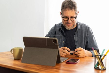A senior man making an online payment using a credit card and tablet, smiling while working from home - Mature man shopping online - Digital transaction, indoor setting, casual atmosphere.