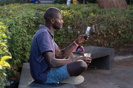 Un joven afroamericano sentado al aire libre en un parque, usando un teléfono inteligente y sosteniendo una bebida - Escena casual y relajada - Tecnología moderna, tiempo libre, fondo natural.