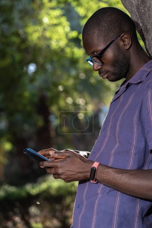 A young African American man using a smartphone while leaning against a tree, wearing glasses and a casual shirt in a park setting - Casual and modern technology use - Green and natural background.