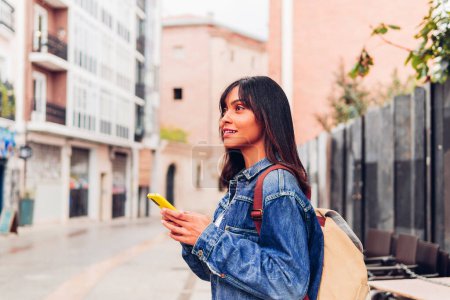 Photo for Side view of young Hispanic female in denim jacket with backpack standing on street navigating with mobile phone in hands and looking up at buildings - Royalty Free Image