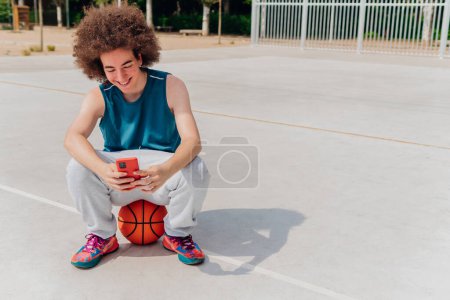 Téléchargez les photos : Photo avec espace de copie d'un adolescent caucasien utilisant le téléphone dans un terrain de basket-ball - en image libre de droit