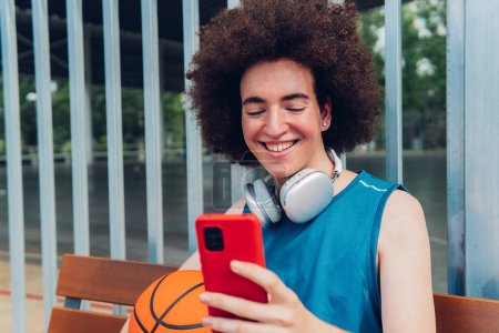 Téléchargez les photos : Garçon utilisant téléphone assis sur un banc d'un terrain de basket-ball urbain - en image libre de droit