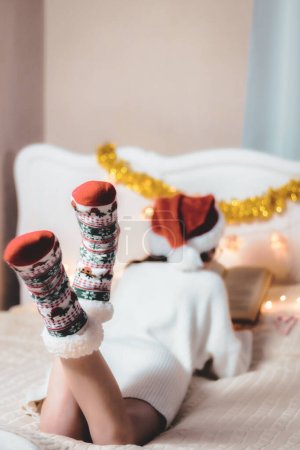 Photo for Caucasian teenage girl in a santa claus hat, in a white knitted dress reads a book lying on her stomach, lifting her legs in socks up on the bed in the bedroom, close-up side view with depth of field. - Royalty Free Image