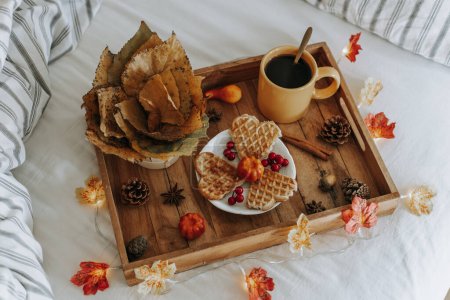 Foto de Una bandeja de madera con una taza de café, gofres en un platillo, hojas de álamo en una cesta de mimbre, decoración de otoño y una guirnalda ardiente acostada en una cama enderezada, vista superior de cerca con profundidad de campo. Concepto de vacaciones de otoño. - Imagen libre de derechos