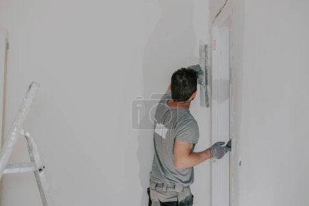Un hombre caucásico joven guapo con un uniforme gris y guantes de pie desde la parte posterior sostiene una pequeña espátula en su mano y masilla un gran ángulo de la puerta, vista lateral de cerca. Concepto de construcción.