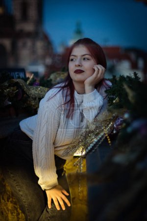 Foto de Retrato de una hermosa adolescente caucásica joven en un suéter de punto blanco con el pelo teñido y los labios mira hacia otro lado con una sonrisa, de pie apoyado en una barandilla festivamente decorada en un balcón - Imagen libre de derechos