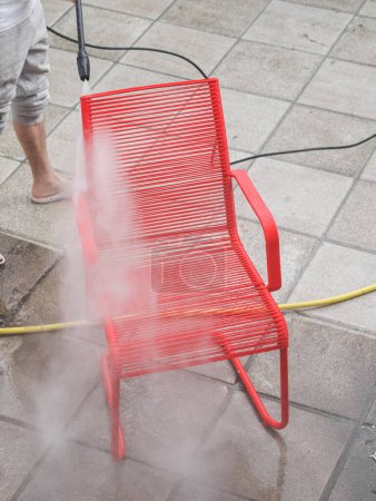 Foto de Un tipo caucásico en pantalones de chándal grises lava una silla de alambre rojo bajo la presión del agua Karcher, que se encuentra en baldosas de piedra en el patio trasero de su casa, vista lateral de cerca. El concepto de lavado de sol - Imagen libre de derechos