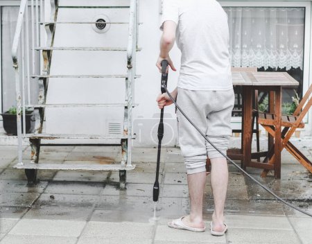 Foto de Un tipo caucásico con una camiseta gris y pantalones de chándal de la parte trasera lava la terraza en el patio trasero de su casa con un karcher, vista lateral, primer plano. El concepto de lavado de tumbonas, limpieza de la casa - Imagen libre de derechos