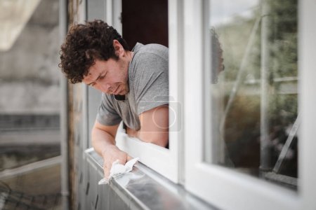 Photo for One handsome caucasian young man with curly brown hair and a smile wipes a window frame with a paper napkin outside while standing in a room being renovated, side view close-up with selective focus. - Royalty Free Image