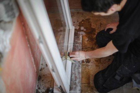Photo for One handsome Caucasian young man with brown hair washes a window frame inside with a sponge and soap while sitting on his knees in a room being renovated, side view close-up with selective focus. - Royalty Free Image