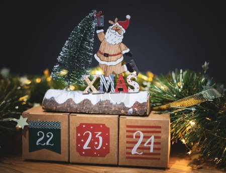 Foto de Hermosas tres cajas kraft calendario de adviento con números 22,23,24 en una mesa de madera con decoración de Navidad y una guirnalda ardiente sobre un fondo oscuro borroso, vista lateral de cerca. - Imagen libre de derechos