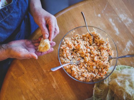 Hands of a senior woman wrapping a raw sauerkraut stuffed cabbage stuffed with rice and meat stuffing holding it in her hand, sitting at a round table in the kitchen, close-up top view. The concept of step by step instructions, home cooking, traditio