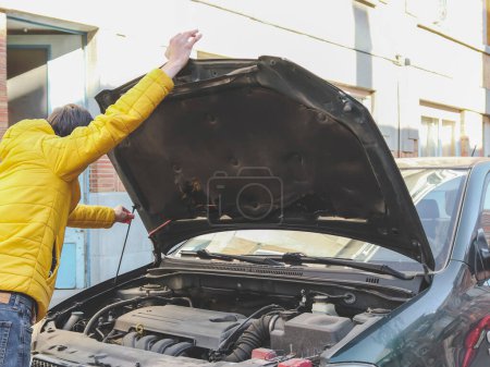 A young caucasian guy in a yellow jacket holds the open hood of a car with one hand, and fastens it with the other to check the breakdown of the engine of his car on a city street in front of the house, close-up side view. Home repair engine concept.