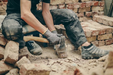 Photo pour A young caucasian man in black work clothes sits on a small chair on the left and cleans the bricks from dirt, debris and old cement with an ax, side view close-up.Construction work concept. - image libre de droit