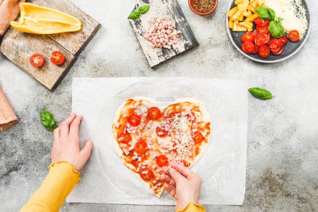 Téléchargez les photos : Des mains d'adolescente caucasienne déposent une tomate cerise tranchée sur une pâte à pizza en forme de cœur pour la Saint-Valentin avec des ingrédients sur la table, vue latérale rapprochée. Valentine's day pizza making concept. - en image libre de droit