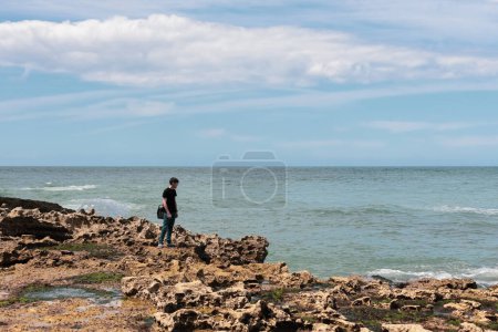 Téléchargez les photos : A handsome caucasian young brunette guy in jeans and a t-shirt stands on a rocky shore looking at the north sea in Etretat in France, close-up view from above. People and nature concept. - en image libre de droit